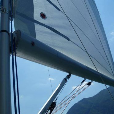 Albero e vela di una barca a vela contro un cielo azzurro brillante, con montagne visibili sullo sfondo.
