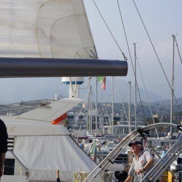 Un uomo su una barca a vela sistema l'attrezzatura in un porto, con altre barche e montagne sullo sfondo.