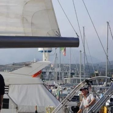 Albero e vela di una barca a vela nel porto con barche e persone.