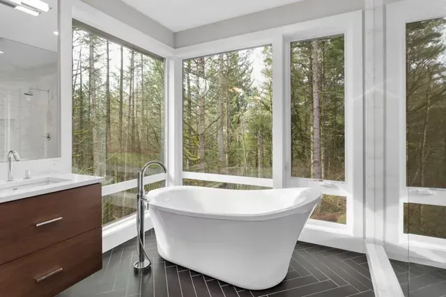 Bathroom with a white freestanding tub, large windows overlooking a forest, dark floor, and wooden vanity.