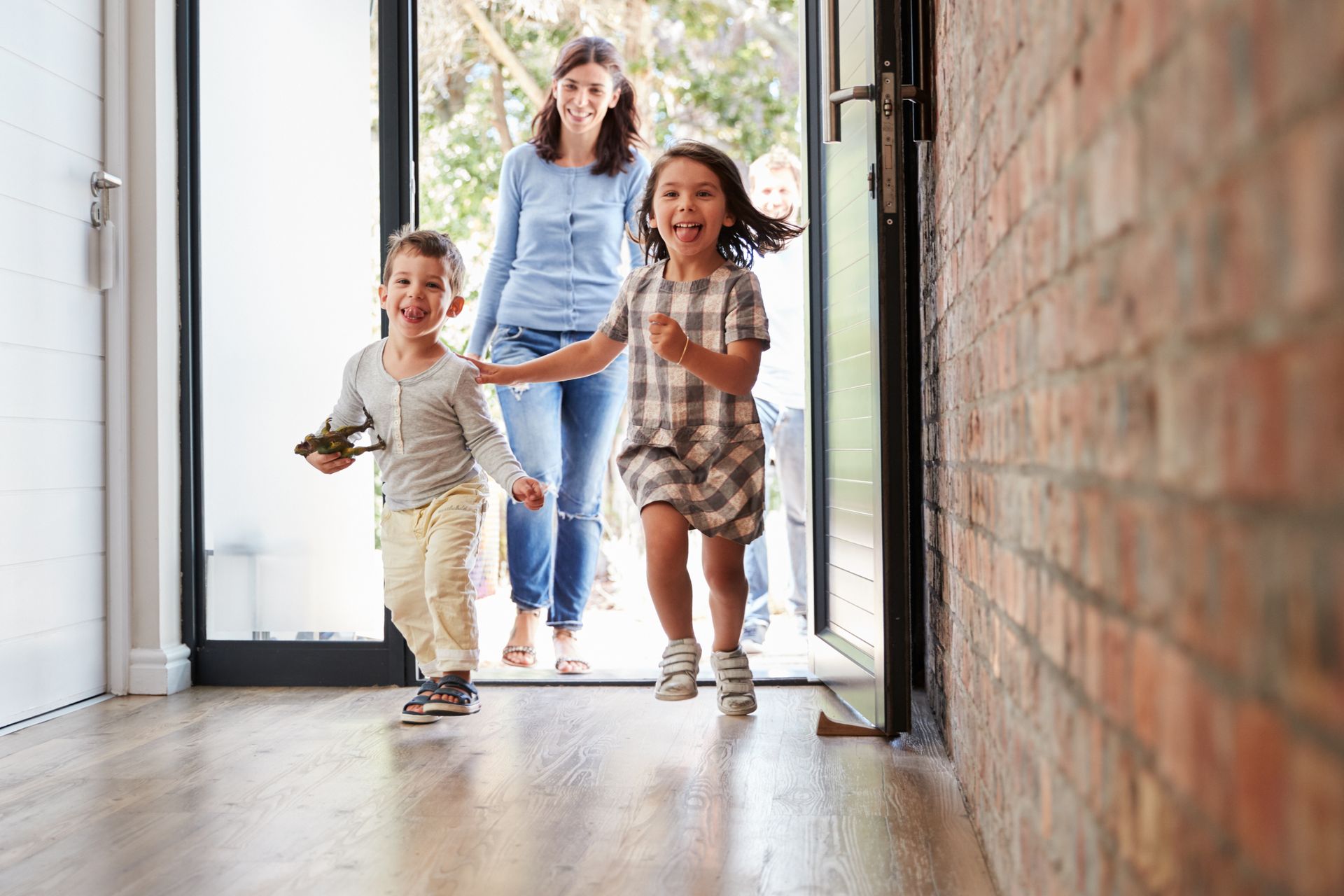 Woman watches two children run toward the camera, doorway open. Brick wall on right.