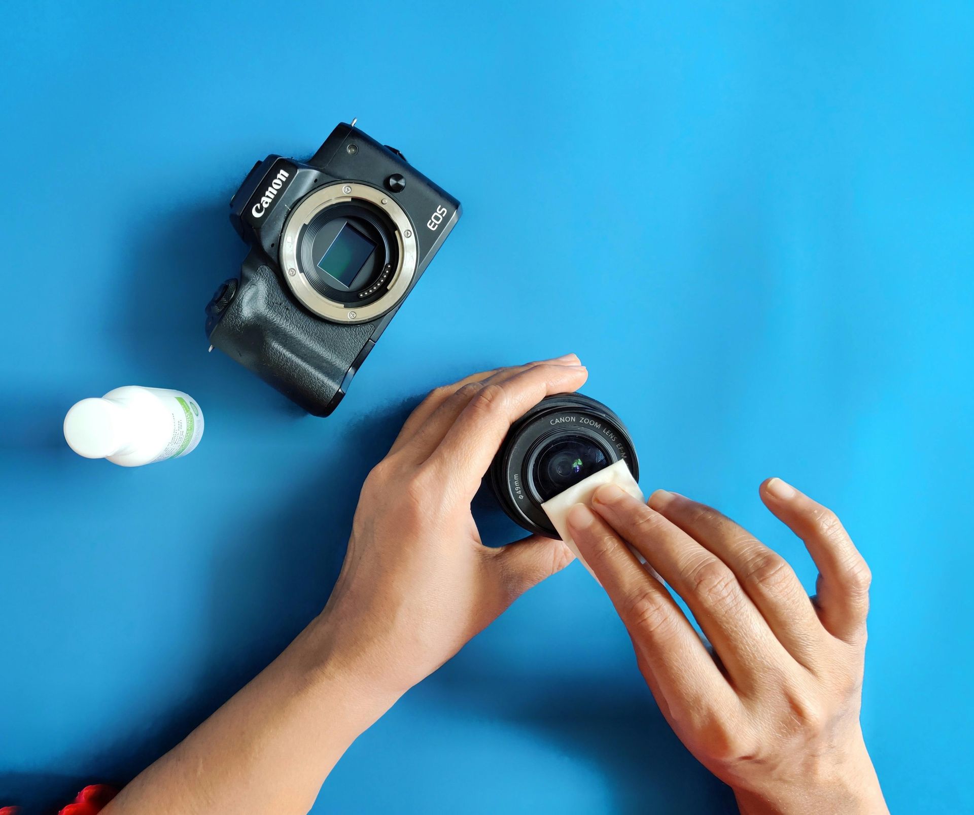 Hands cleaning a camera lens with a wipe; a camera body and cleaning solution are also on a blue surface.