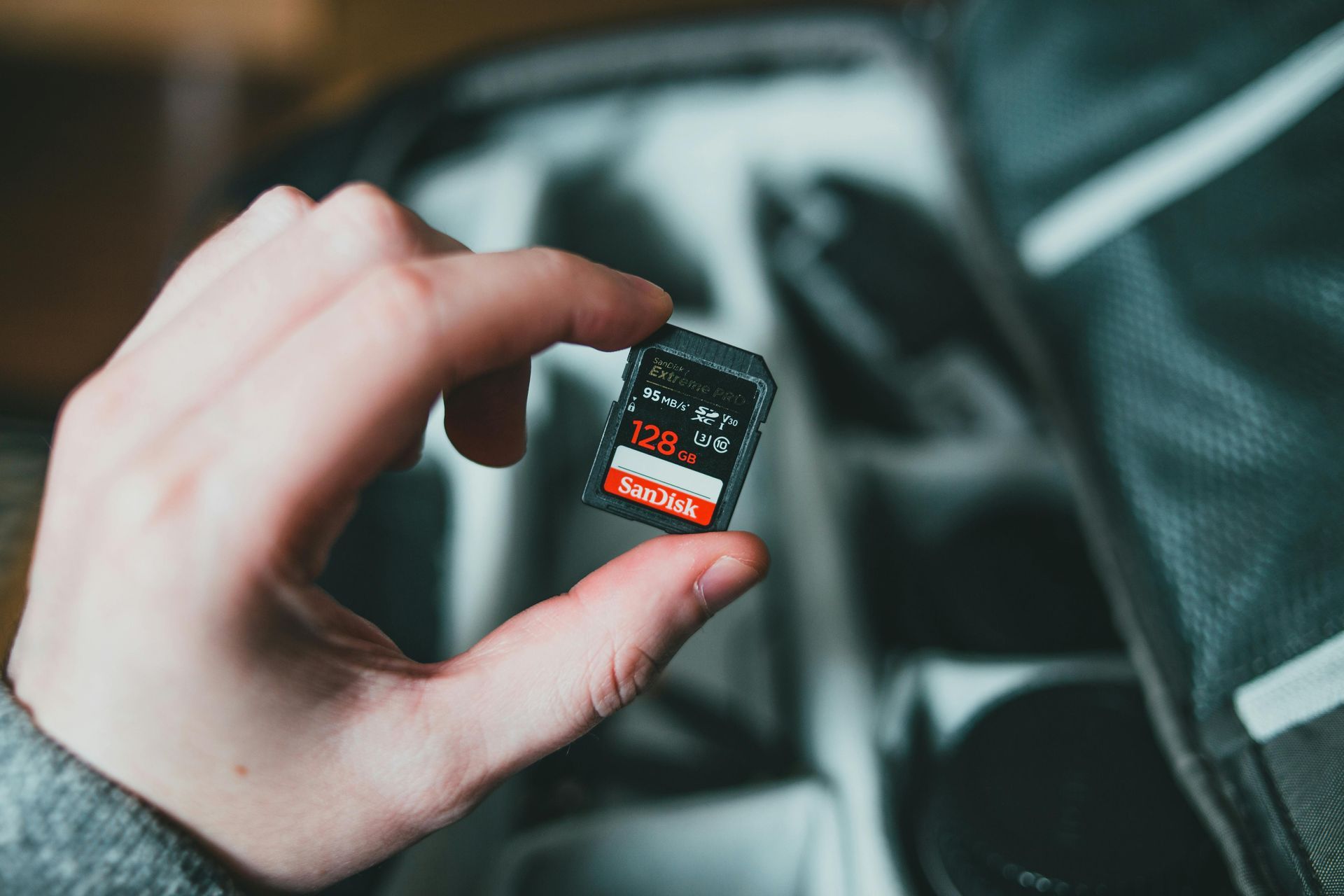 Hand holding a small black and red SD card, with a blurred camera bag in the background.