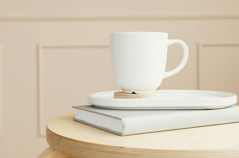 White cup on stacked books and a plate on a light wood table against a beige wall.