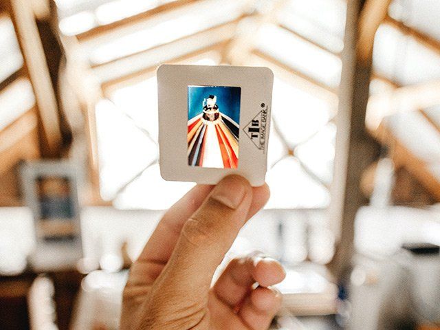 Hand holding a slide with an image of a person with multicolored robes, set against a window backdrop.