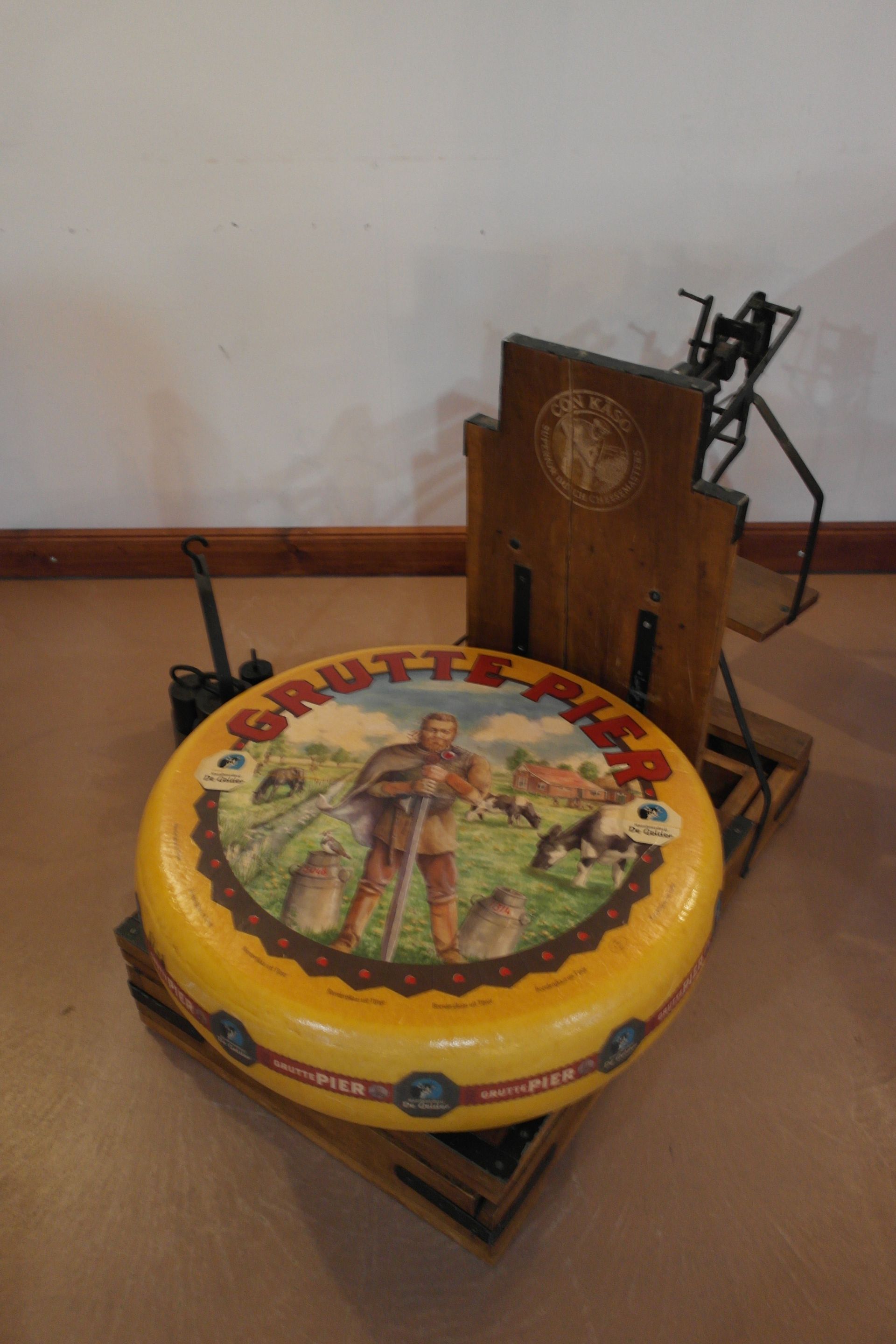 Runder Käselaib auf einem Holzsockel mit Käseschneider; das Etikett zeigt eine Person auf einem Feld.