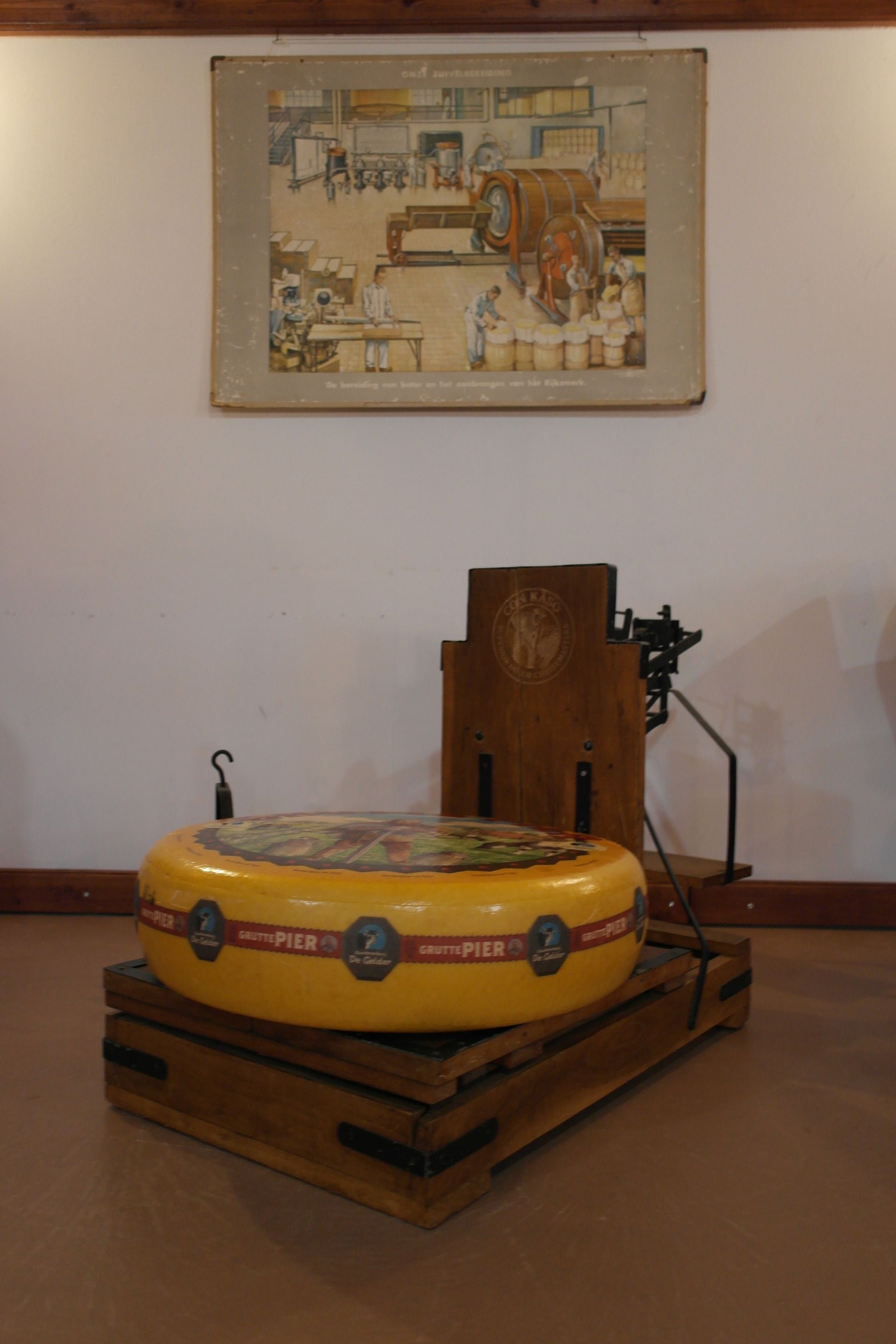 Ein großer gelber Käselaib auf einem Holzständer, im Hintergrund eine Skala. Darüber hängt eine Zeichnung.