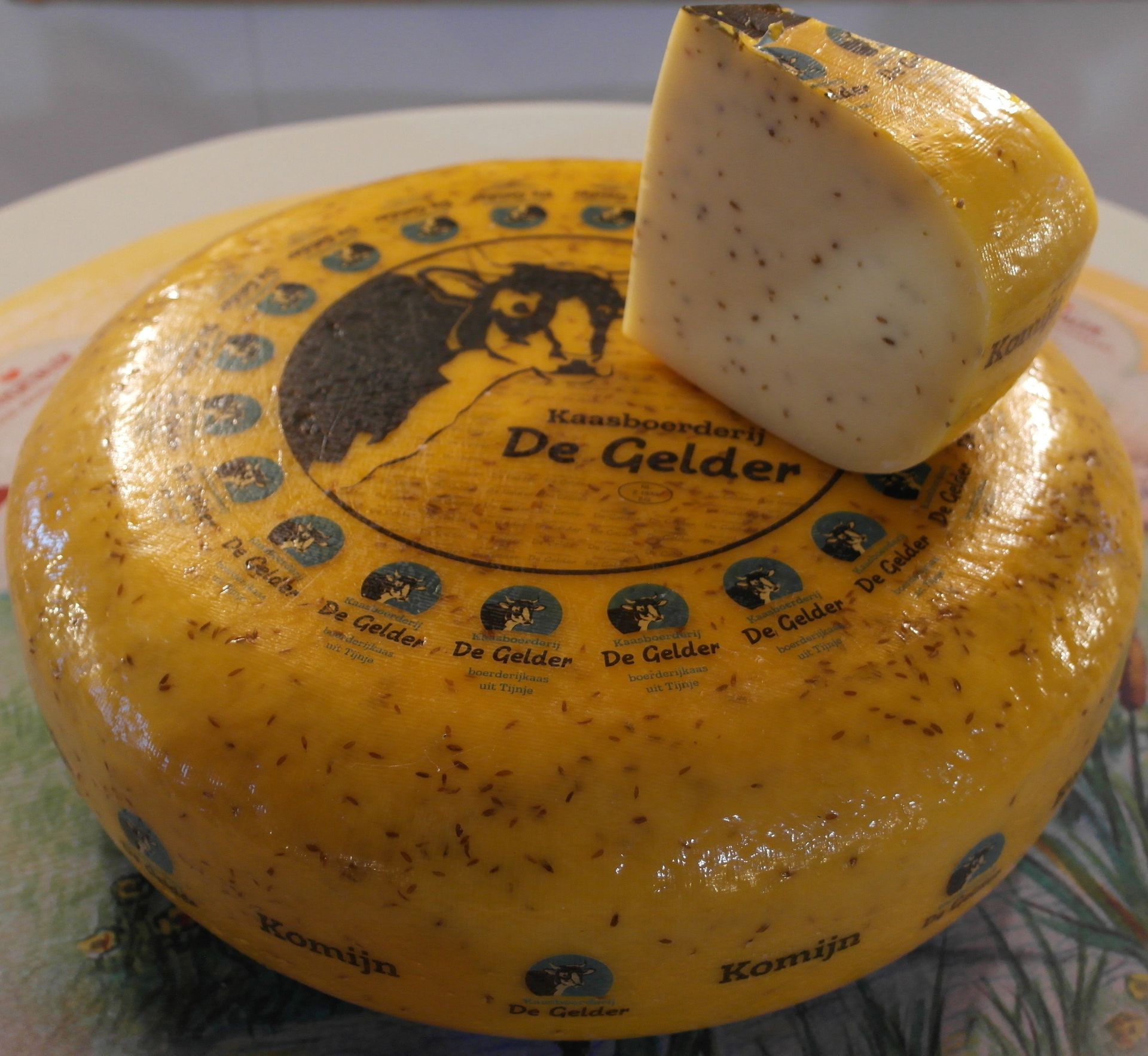Runder Käse „De Gelder“ mit angeschnittenem Keil, gelber Rinde und Logo obenauf.