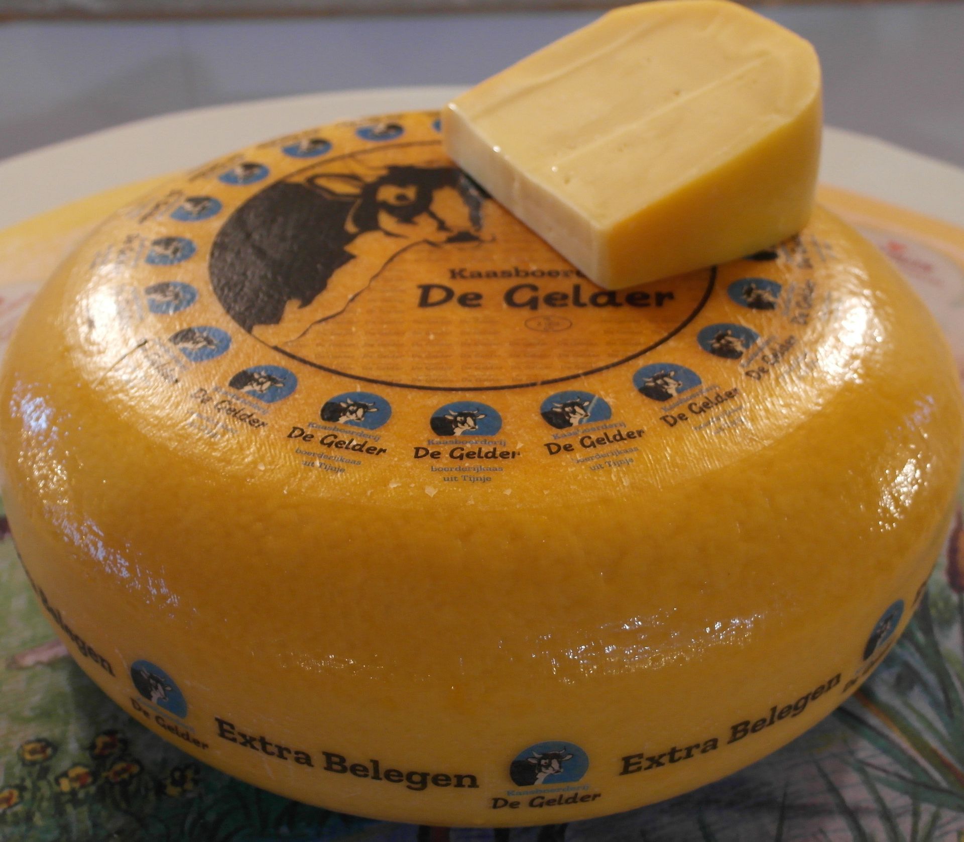 Ein Laib holländischer Käse, „De Gelaer“, mit einem angeschnittenen Stück. Gelbe Wachsrinde, blaues Etikett.