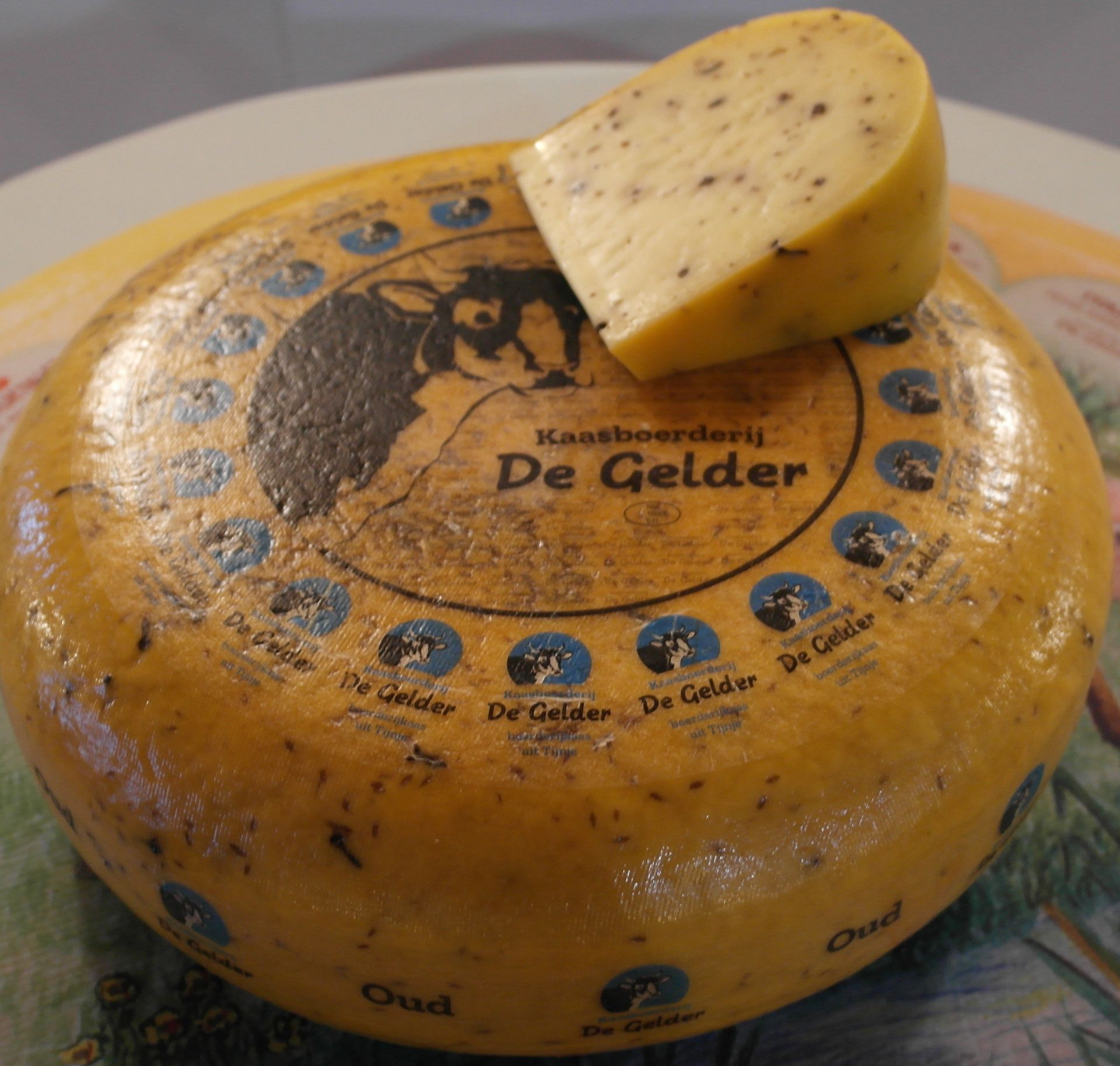 Runder Käselaib von De Gelder mit einem Keil obendrauf; gelb mit dunklen Sprenkeln und Markenzeichen.