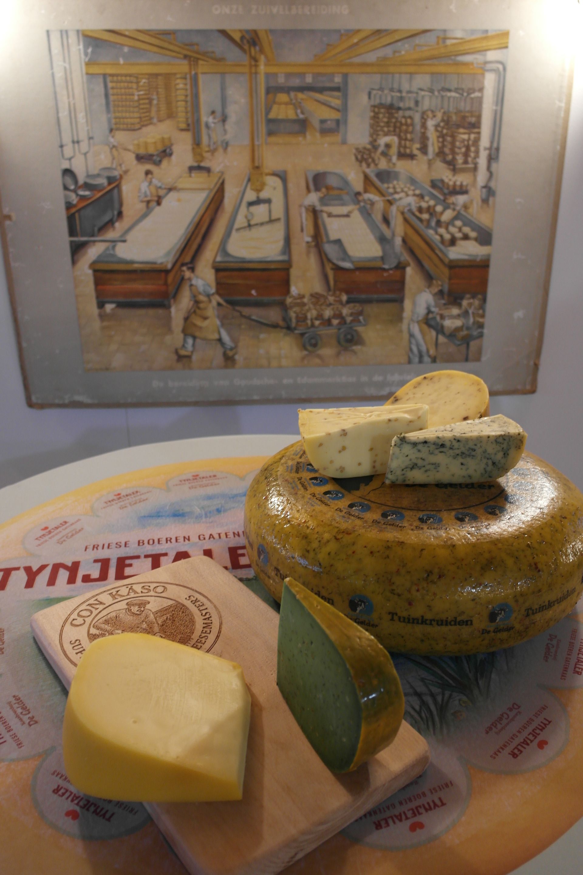 Verschiedene Käsesorten sind auf einem Tisch; Hintergrund ist ein Gemälde einer Fabrikinneneinrichtung zu sehen.