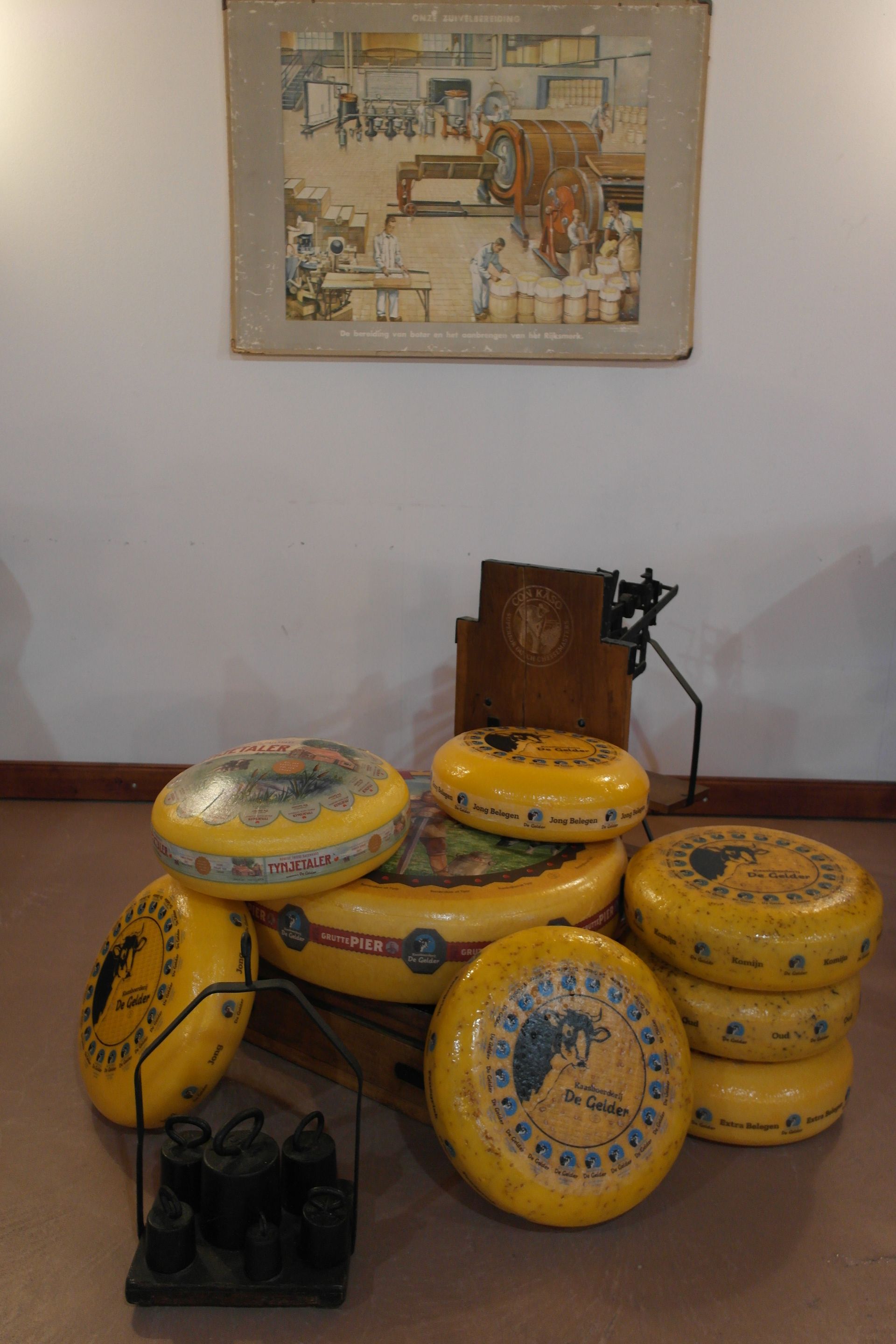 In einem Zimmer stehen gestapelte, gelbe Käseräder unter einer gerahmten Illustration.