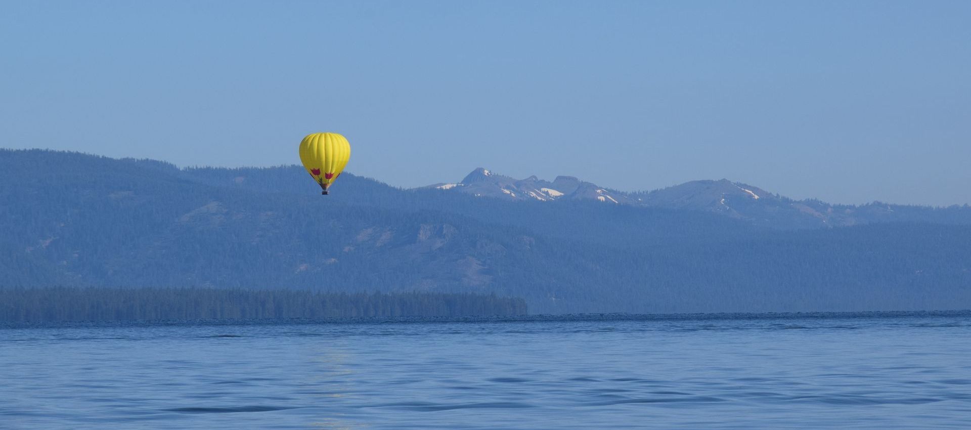 A yellow hot air balloon is flying over a lake with mountains in the background.