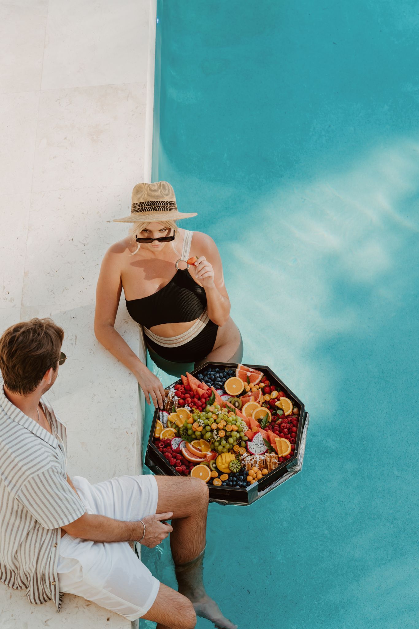 a man and a woman are sitting in a swimming pool with a tray of fruit