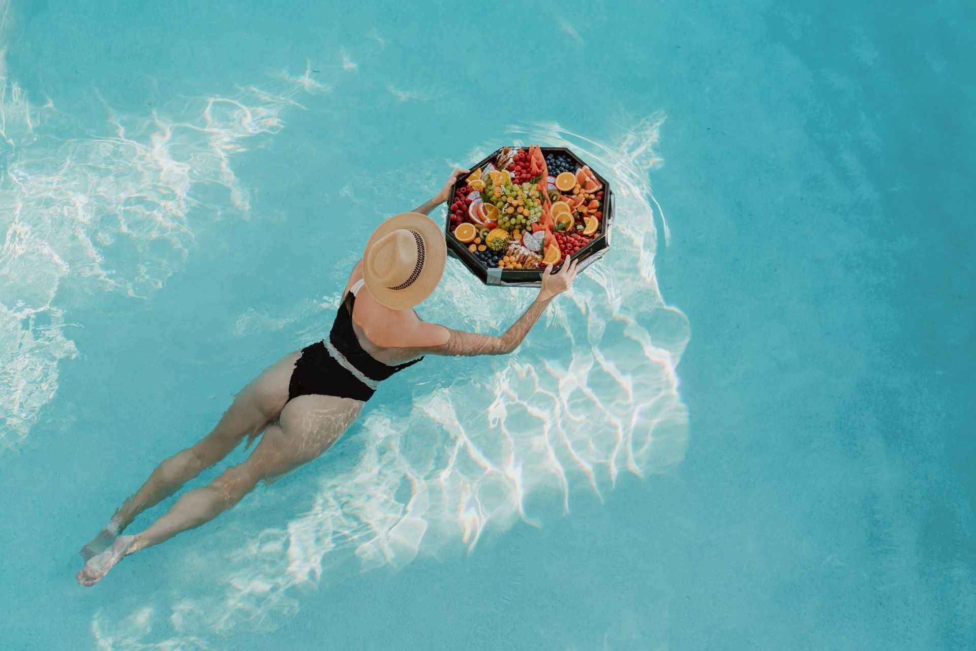 a woman is swimming in a pool holding a tray of fruit