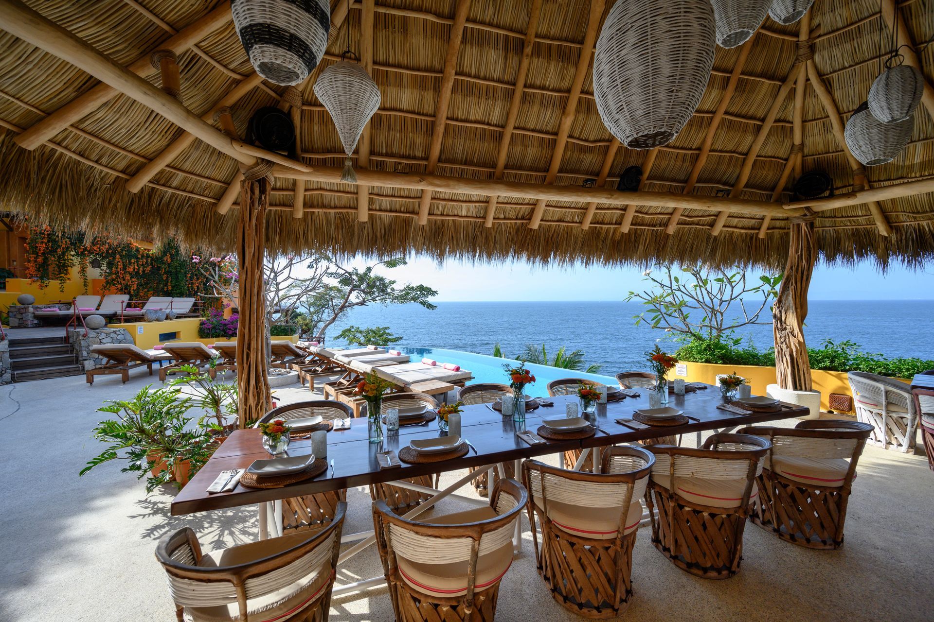 a dining room with a table and chairs under a thatched roof overlooking the ocean