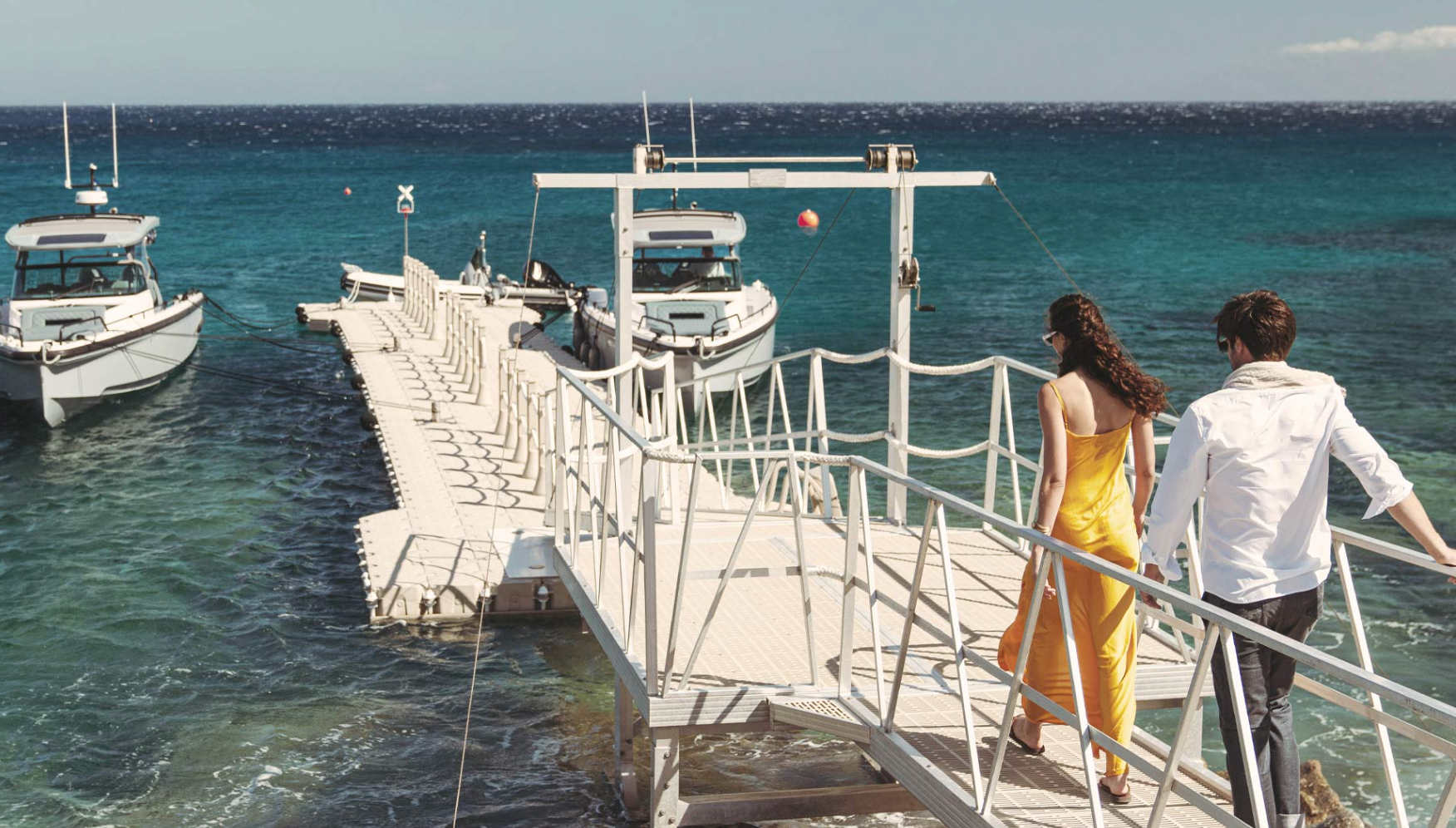 a man and a woman are walking on a dock with boats in the background