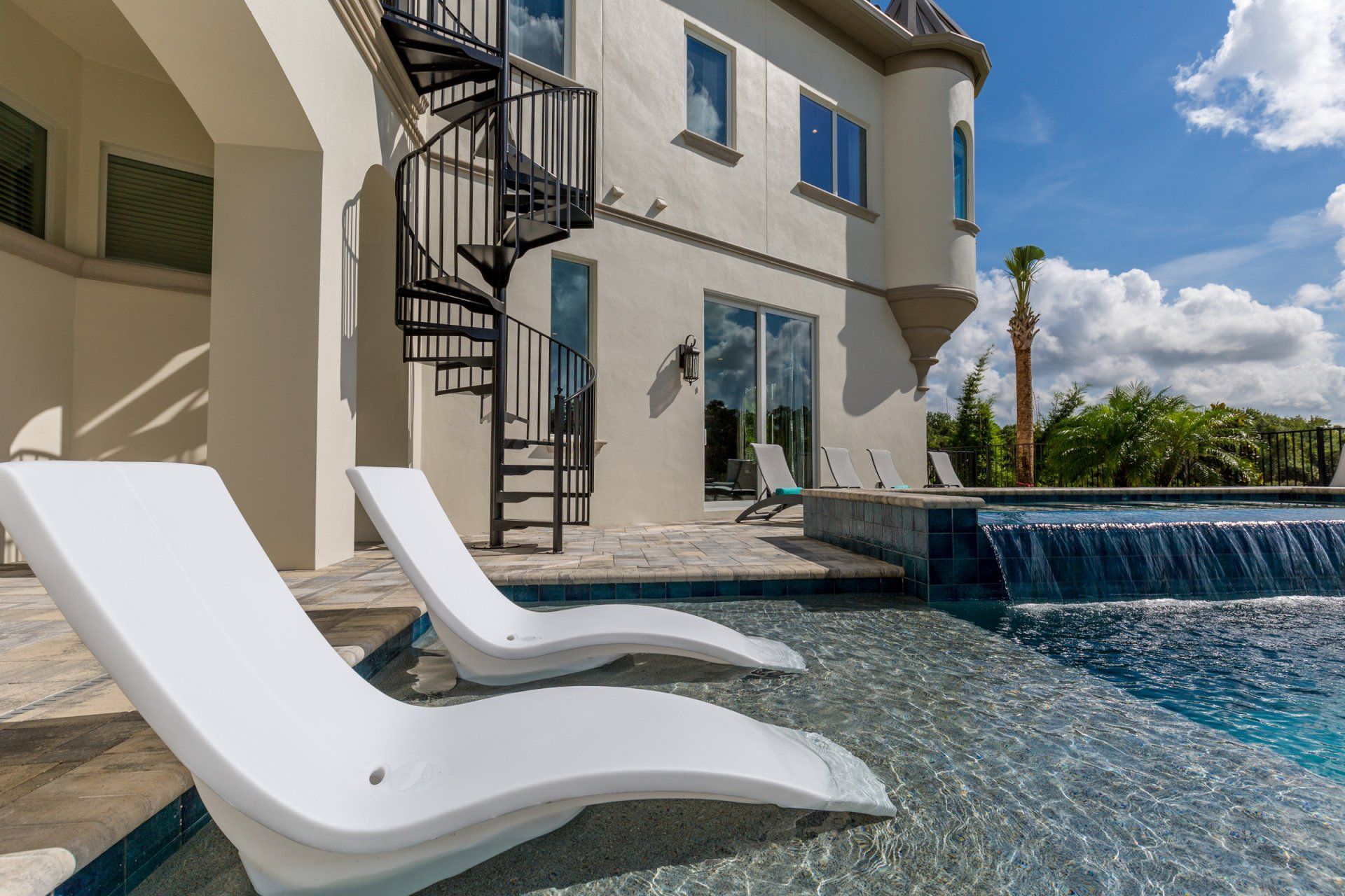 two white lounge chairs are sitting next to a swimming pool in front of a house with a spiral staircase