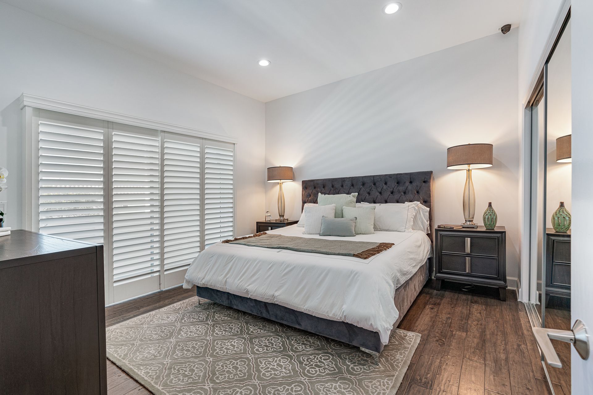 A bedroom with a king size bed , nightstands , lamps and shutters.