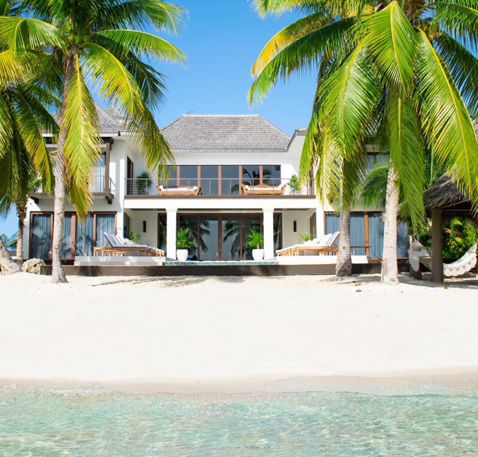 A large white house sits on a beach surrounded by palm trees