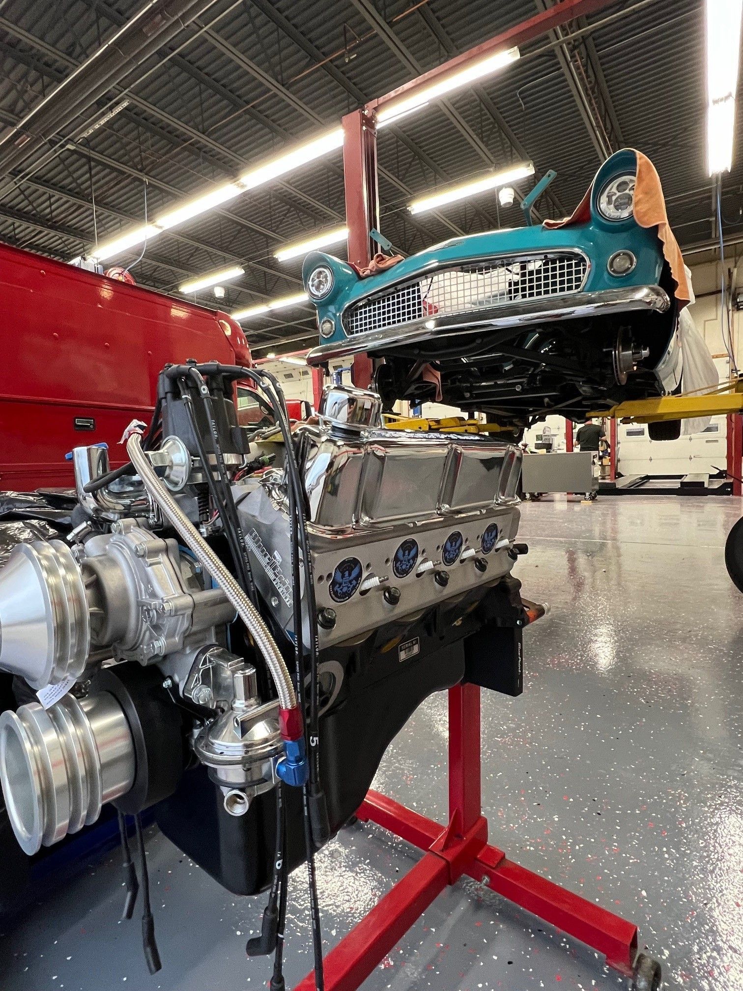 A vintage car front-end, turquoise, being placed atop a large engine on a red stand in a garage.