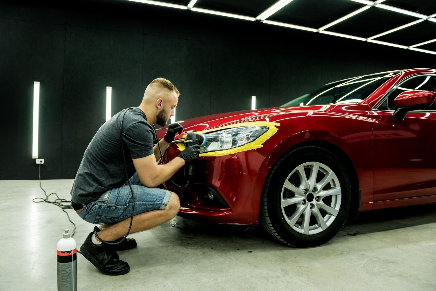 Detailer polishing the front fender of a red car in a dim garage, with a spray bottle nearby