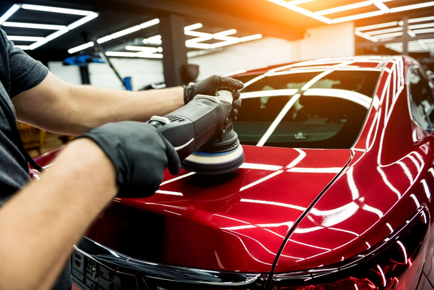 Detailing worker polishing a red car hood in a garage with bright overhead lights