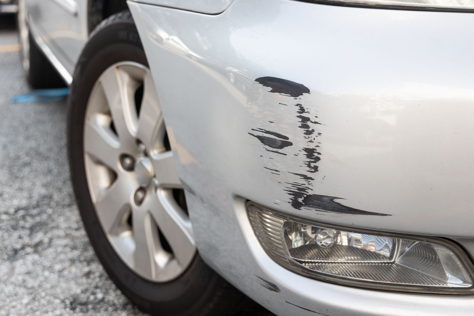 A silver car with a damaged bumper is parked on the side of the road.