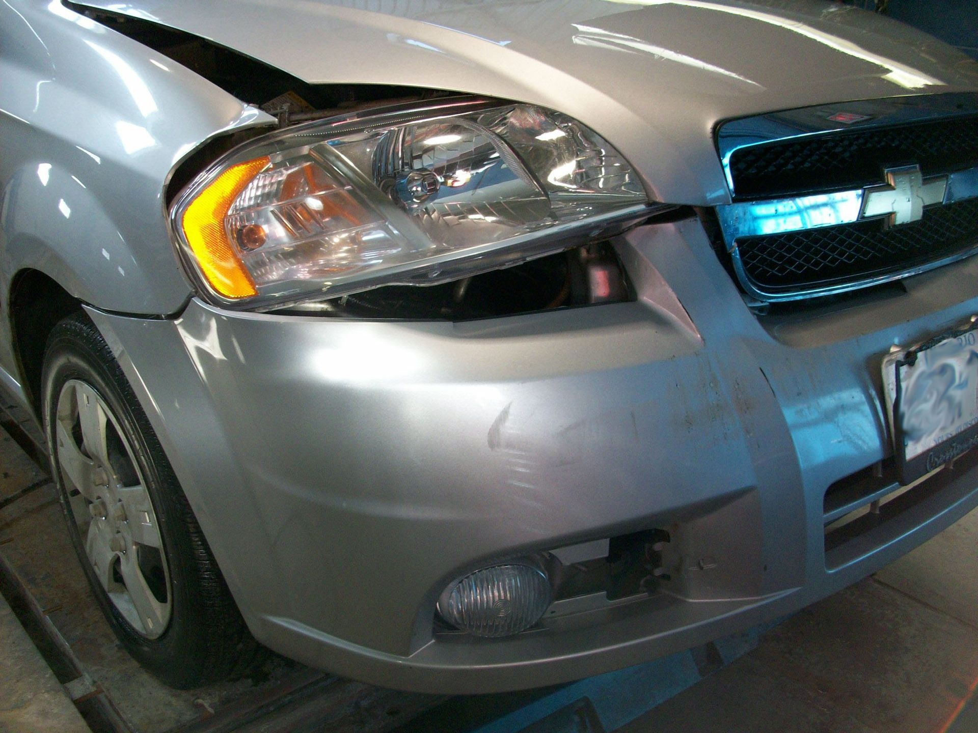 A silver chevy with a broken headlight is on a lift