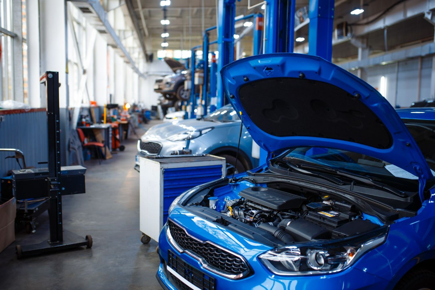 Blue car with hood open in an auto repair shop, showing the engine bay alongside another car on a lift