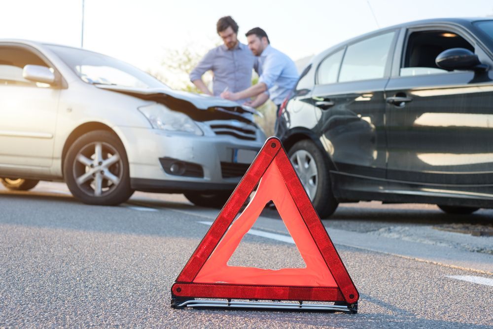 Two men assessing damage to cars after a collision, with a warning triangle in the foreground.