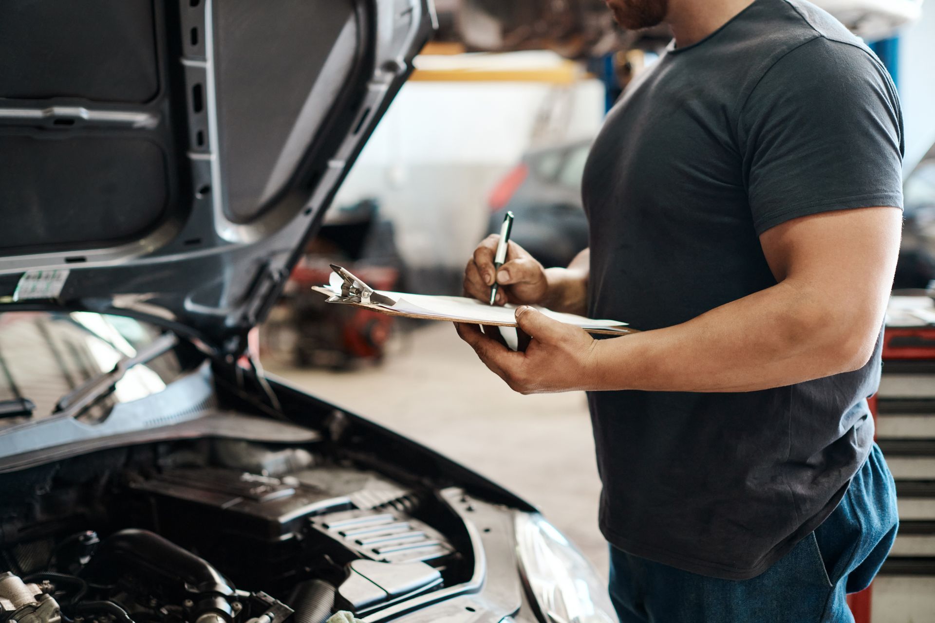 A man is writing on a clipboard while looking under the hood of a car.