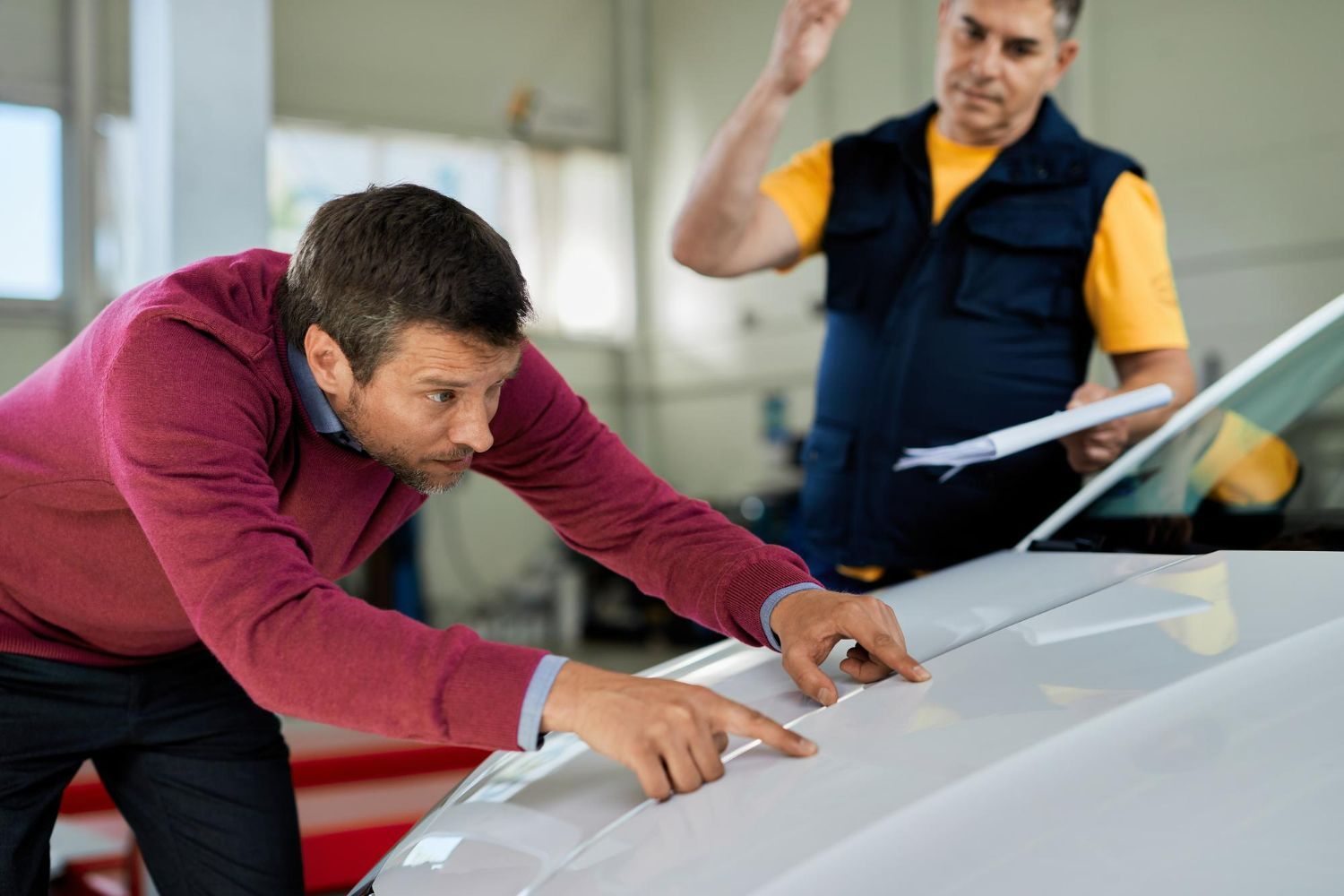Man inspecting car's hood in a garage. Mechanic gestures with papers.