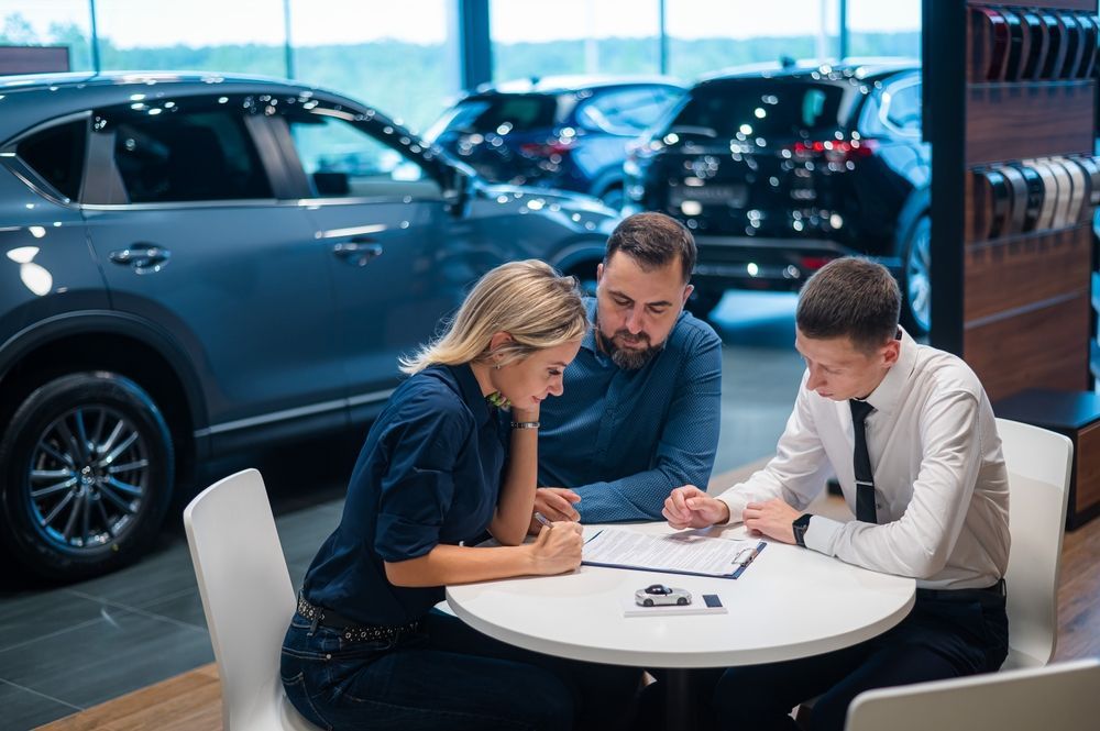 Couple reviews paperwork with salesperson at a car dealership; cars in background.