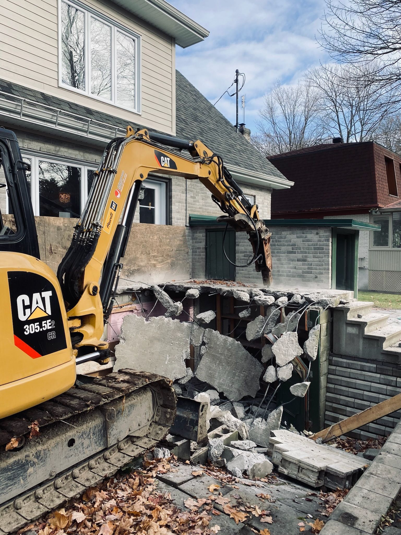 Une excavatrice à chat démolit un mur de briques devant une maison.