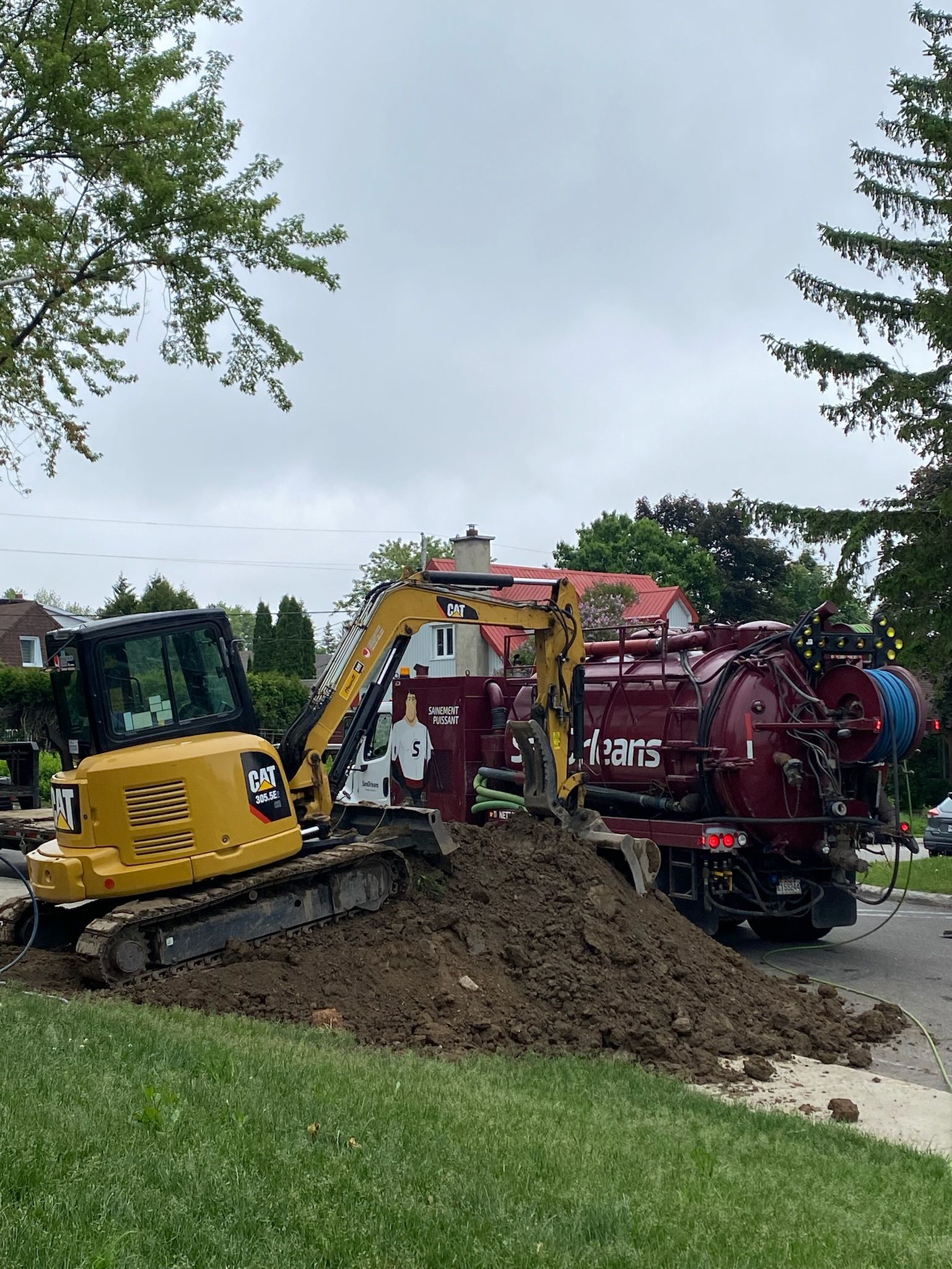Une excavatrice jaune creuse un trou dans le sol à côté d'un camion aspirateur rouge.