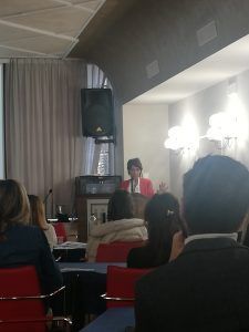 Una donna con una giacca rossa parla sul podio, di fronte al pubblico seduto in una sala conferenze.