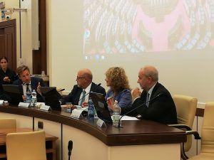 Gruppo di quattro persone sedute a un tavolo curvo, intente a discutere. Sullo sfondo c'è uno schermo per proiettore.