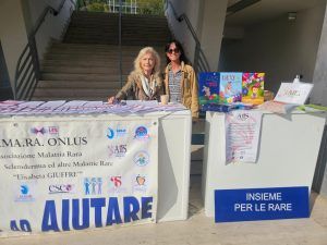 Due donne sedute a un tavolo con opuscoli e libri, promuovono un gruppo di sensibilizzazione sulle malattie rare, 