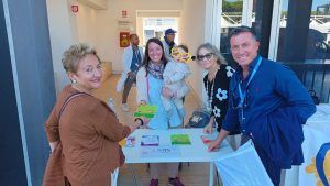 Persone sedute a un tavolo che distribuiscono materiali. Donna che tiene in braccio un bambino, ambiente soleggiato all'aperto.