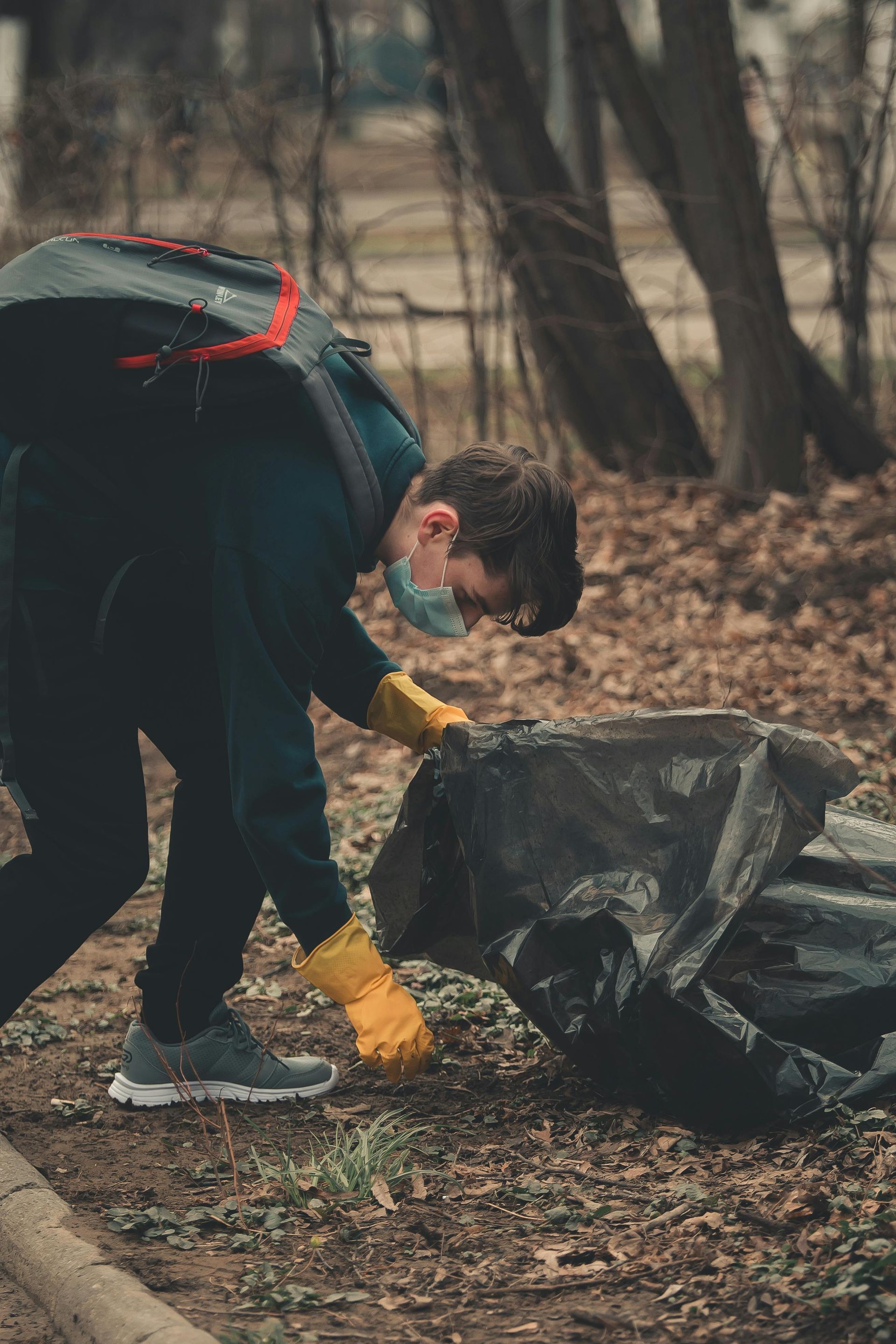 Person wearing a mask and gloves picking up trash, filling a black bag in an outdoor setting.