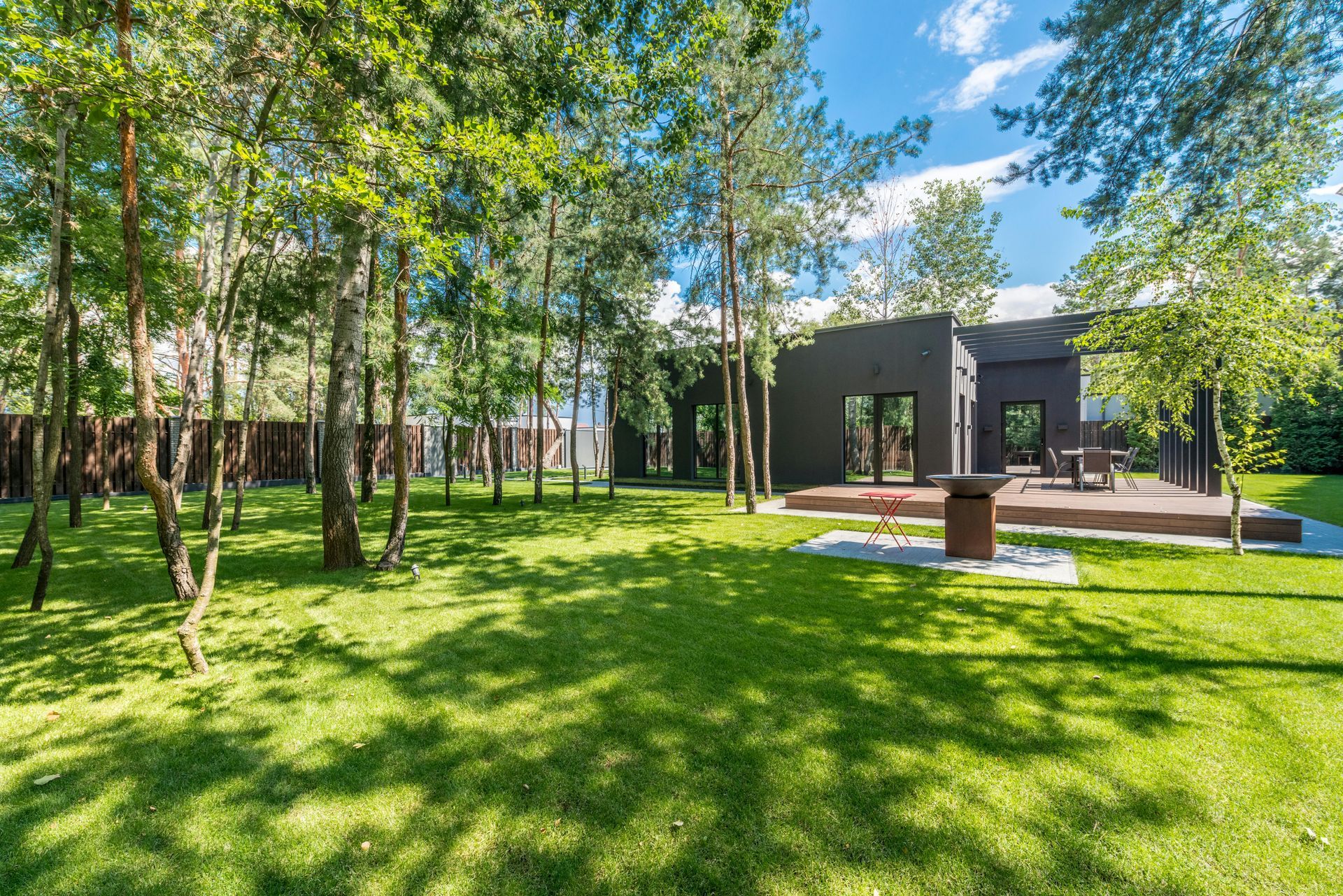 Black house with wooden deck, surrounded by green grass and trees on a sunny day.