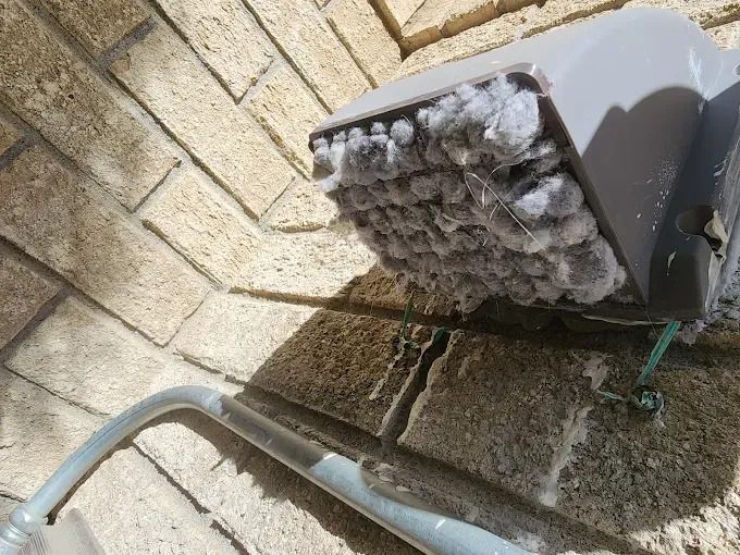 Outdoor utility box heavily clogged with gray lint or debris beside a brick wall.