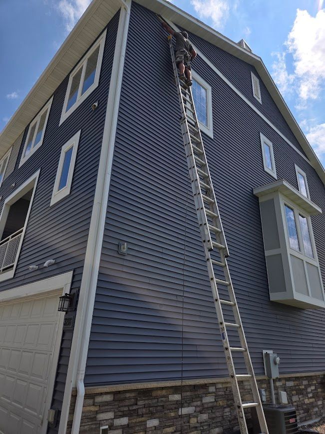 Person on a tall ladder, working on the blue siding of a two-story house with white trim, on a sunny day.