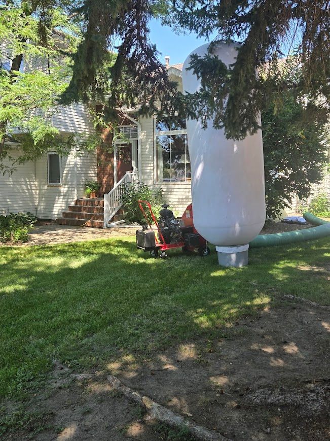 White house with a large white cylindrical container. A machine and green hose are near it, on a grassy lawn.