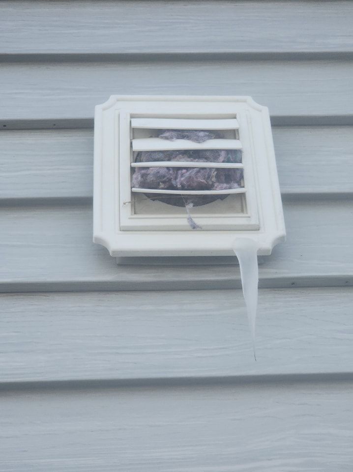 White dryer vent on gray siding, blocked with a dark mass and an icicle below.