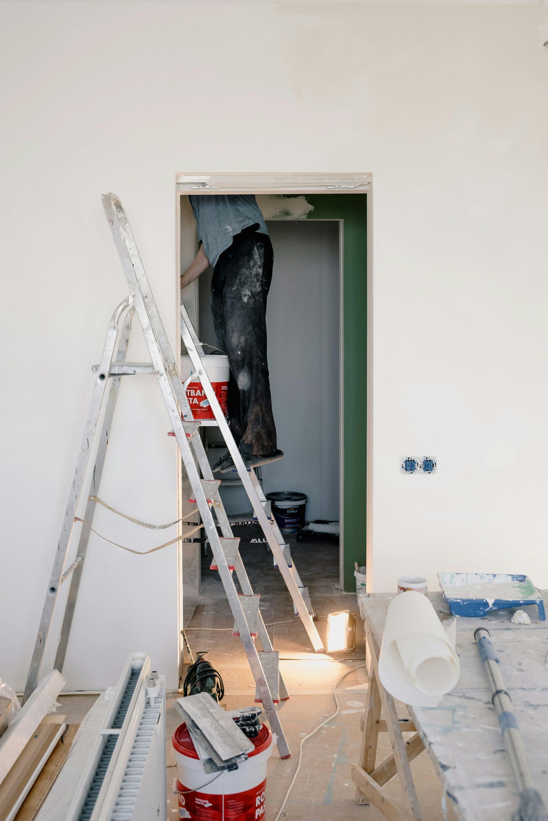 Renovation in progress: A person in doorway with ladders, paint buckets, and tools. White walls, green door frame.
