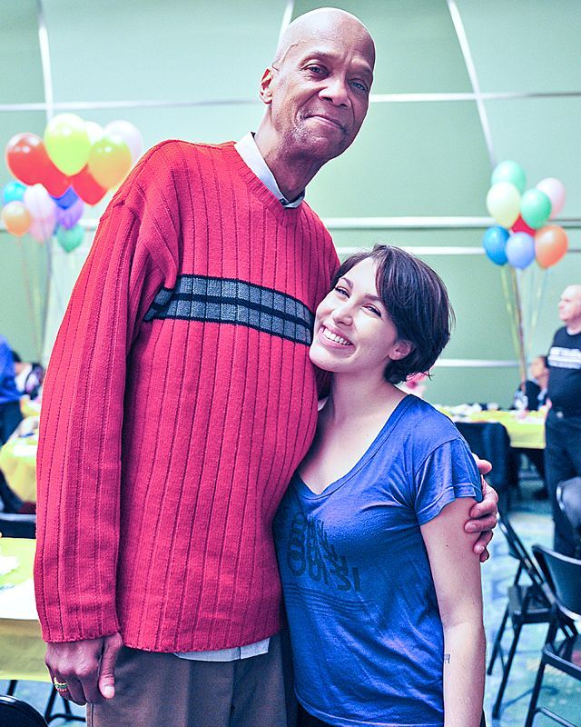 Man in red sweater hugs woman in blue shirt, smiling at a gathering with balloons.