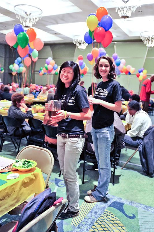 Two people, wearing matching black shirts, smile and stand in a room decorated with balloons.