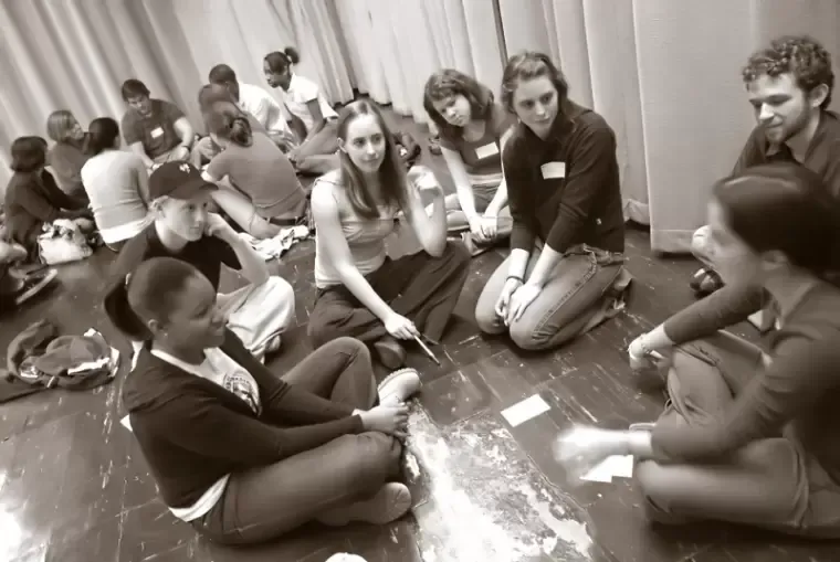 People in a circle on floor, in a room. Some are talking, holding objects.